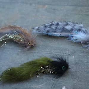 May include: Three fly fishing lures with different colored feathers and bodies. The lures are brown, gray, and black with green accents.