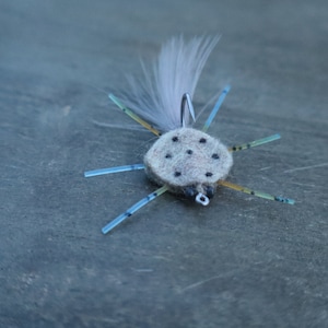 May include: A gray and white fly fishing lure with a white feather tail and six blue and yellow legs.