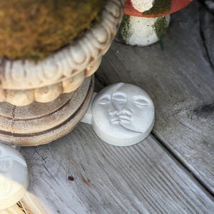 May include: A light-colored, round, stone garden accent featuring a crescent moon and face design. The moon-shaped accent is placed on a wooden surface near a decorative planter and a mushroom-shaped garden ornament.