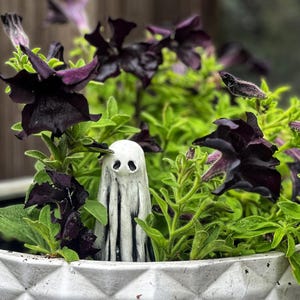 May include: A white ceramic ghost figurine with black eyes and a striped pattern is nestled in a pot of dark purple petunias and green foliage.