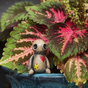 May include: A small, beige, textured figurine resembling a doll with large black button eyes and a stitched mouth. It features a bright red heart patch on its chest and a small yellow patch on its lower body, sitting upright with rounded limbs.