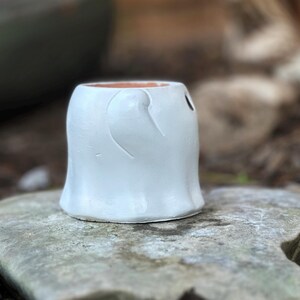 May include: A handmade white ceramic ghost-shaped planter. The planter has a rounded body with a curved design on the front and a small hole for a face. The interior is a warm brown color. The planter is sitting on a gray stone.