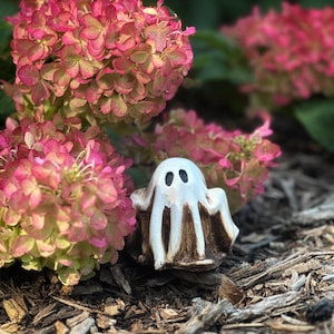 May include: A white ceramic ghost figurine with black eyes, sitting on a bed of brown wood chips and surrounded by pink hydrangea flowers.