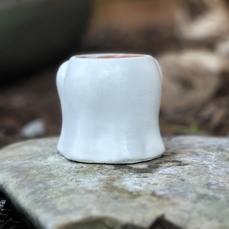 May include: A small, white ceramic mug with a rounded body and a small handle. The interior of the mug is a warm, earthy brown. The mug is sitting on a gray stone surface, with a blurred natural background.