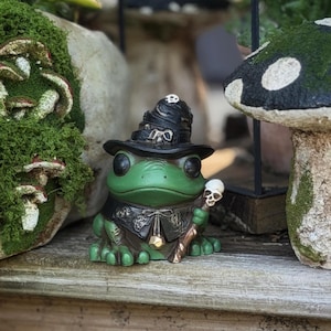 May include: A green frog figurine wearing a black witch hat with skull accents and a matching cloak. The frog holds a staff topped with a skull. The figurine is a whimsical garden decoration.