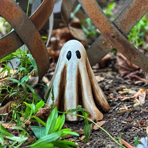 May include: A white ceramic ghost figurine with black eyes, sitting in a garden setting with green plants and brown dirt.