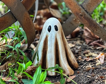 Può includere: Una statuetta di fantasma in ceramica bianca con occhi neri, seduta in un giardino con piante verdi e terra marrone.