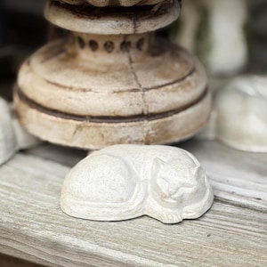 May include: A small, off-white cat figurine in a sleeping pose. The cat is sculpted with simple lines and details, resting on a wooden surface. The background includes a decorative, aged wooden object.