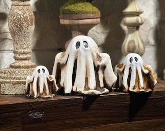 Può includere: Tre figurine di fantasmi in ceramica bianca con occhi neri. I fantasmi sono di dimensioni diverse e sono disposti in fila su una superficie di legno.