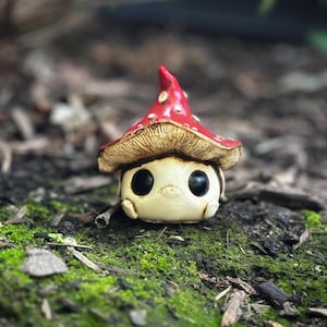 May include: A whimsical ceramic mushroom figurine with a red cap dotted with white spots. The mushroom has a beige stem and a friendly face with large black eyes. The figurine is set on a bed of green moss and earth.