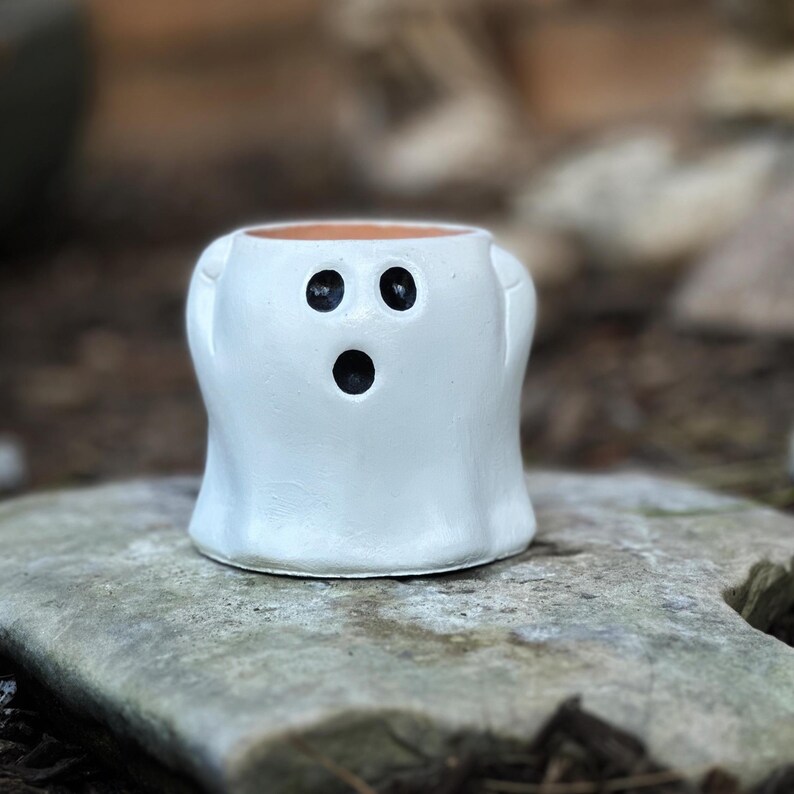 May include: A white ceramic ghost-shaped planter with black eyes and a mouth. The small, handmade pot has a rounded body and two small side extensions. The interior is a natural terracotta color. The planter is sitting on a gray stone.