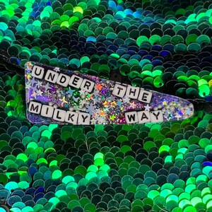 May include: A sparkly purple and green hair clip with the words "UNDER THE MILKY WAY" spelled out in white letters.