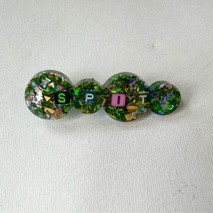 May include: A green resin hair clip with the word "SPOT" spelled out in individual letter beads. The clip is decorated with glitter and small star-shaped confetti. The letters are black and pink.