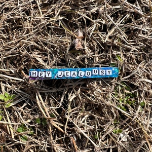 Op de afbeelding: Een rechthoekige kralenarmband met de woorden "HEY JEALOUSY" in witte letterkralen. De armband heeft een blauwe en paarse achtergrond en ligt tegen een achtergrond van droog gras en kleine groene planten.