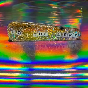 May include: A clear plastic hair clip with gold glitter and white letter tiles spelling "TO THE LEFT".