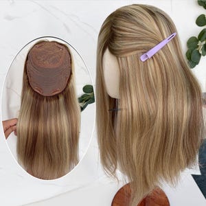 May include: A hair topper with straight, highlighted blonde and light brown hair. The piece features a brown lace base with integrated clips for attachment. A light purple hair clip is pinned to the side of the hair on a mannequin head.