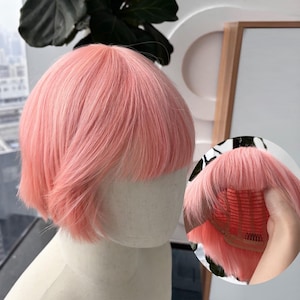 May include: A short, pink wig with a blunt fringe, styled on a mannequin head. The wig has a side part and a slightly angled cut. A close-up shows the wig's cap and construction.