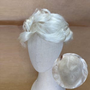 Pode incluir: Uma peruca loira clara com um estilo curto e ondulado é exibida em uma cabeça de manequim. A peruca tem uma linha de cabelo de aparência natural e uma aparência macia e texturizada. Um close-up mostra a touca da peruca.