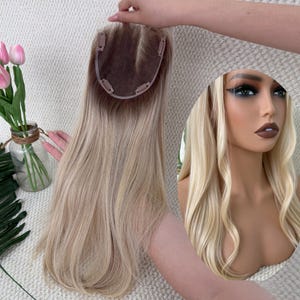 May include: A blonde hair topper with a dark brown base and light blonde highlights. The hairpiece has clips for attachment and is held up by a hand. A mannequin head with long blonde wavy hair is in the background.