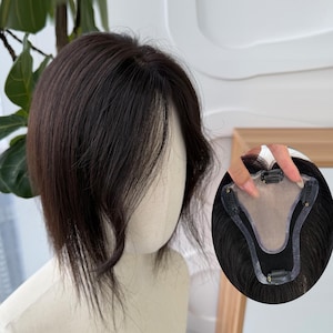 Puede incluir: Una pieza de cabello castaño oscuro con una línea de cabello de aspecto natural. La pieza de cabello está peinada con una raya lateral y ondas suaves. La imagen también muestra un primer plano de la base con clips para una fijación segura.