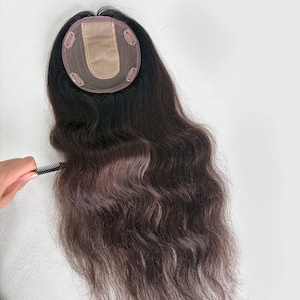 May include: A dark brown hair topper with a light brown and gray ombre effect. The hairpiece has a small, circular base with clips for attachment. A hand is shown combing the hair with a black comb. The hair is wavy and long.
