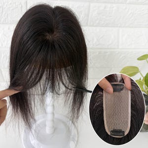 May include: A dark brown hair topper with a centre part and wispy bangs, displayed on a white stand. An inset circular image shows the underside of the hair piece, featuring a beige mesh base with two black metal pressure-sensitive clips.