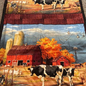 May include: A fabric quilt featuring a pastoral farm scene. The quilt depicts red barns, cows, sheep, pigs, and a windmill under a blue sky with autumn foliage. The quilt has a black border.