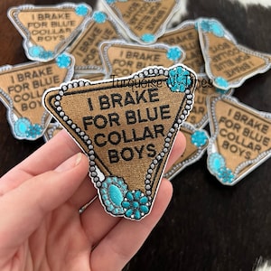 May include: A brown felt patch with a triangle shape and a white embroidered border. The patch has the text "I Brake For Blue Collar Boys" in white embroidery. The patch is decorated with a silver chain and turquoise rhinestones.