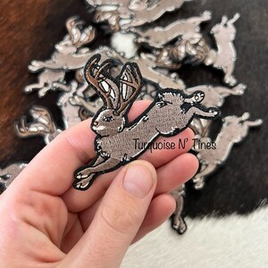 Puede incluir: Un parche bordado marrón y gris de un conejo con cuernos saltando. El parche está sostenido en una mano. El texto "Turquoise N' Tines" es visible en la imagen.