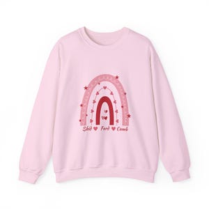 May include: A light pink sweatshirt featuring a rainbow design in shades of pink and red. The rainbow is adorned with hearts and stars, with the words "Shid", "Fard", and "Cumb" below the rainbow.
