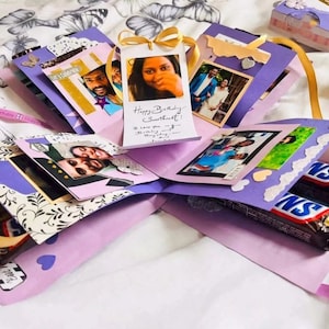 Może przedstawiać: Fioletowe pudełko eksplodujące z fotografiami i ręcznie napisanym komunikatem, który brzmi "Happy Birthday Sweetheart! I love you and I'm celebrating you today!" Pudełko jest ozdobione koronką, sercami i motylami.