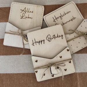 Könnte beinhalten: Kartenhalter aus Holz mit verschiedenen Botschaften. Einer sagt "Alles Liebe", ein anderer "Herzlichen Glückwunsch" und ein dritter "Happy Birthday". Jeder Halter hat eine Tasche und ist mit Bindfaden gebunden. Einige haben herzförmige Verzierungen.