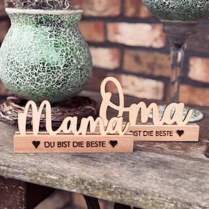 Może przedstawiać: Drewniane znaki dekoracyjne ze słowami "Mama" i "Oma" w kursywie. Każdy znak stoi na małej drewnianej podstawie z napisem "DU BIST DIE BESTE" i symbolem serca. Znaki są jasnobrązowe.