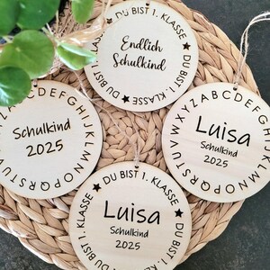 Puede incluir: Cuatro adornos redondos de madera con texto. Uno dice "Endlich Schulkind" y "Du bist 1. Klasse". Dos dicen "Luisa Schulkind 2025". El último dice "Schulkind 2025". Cada uno tiene una cuerda para colgar.