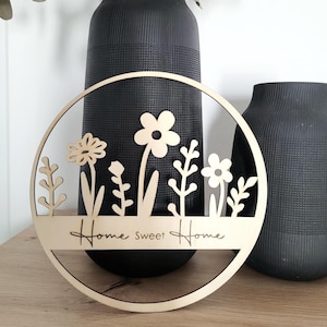 Könnte beinhalten: Runder Wandbehang aus Holz mit floralem Design und dem Schriftzug "Home Sweet Home". Das naturfarbene Holzstück zeigt ausgeschnittene Blumen und Blätter in einem kreisförmigen Rahmen, geeignet für die Heimdekoration.