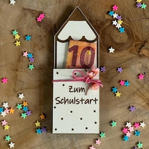 Op de afbeelding: Een houten potloodvormige geschenkdoos met een briefje van tien euro erin, vastgebonden met roze touw en een bloem. De doos heeft de tekst "Zum Schulstart" en is omringd door kleurrijke sterrenconfetti.