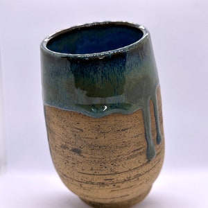 May include: Handmade ceramic tumbler. The base is textured and tan, while the top is glazed in dark blue and green, with drips down the sides. The interior is a deep blue. A unique, artistic piece.