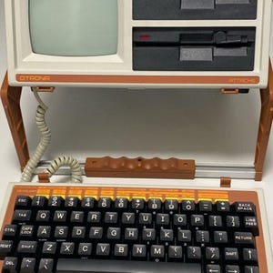 May include: An Otrona Attache portable computer featuring a beige chassis, a small screen, and two disk drives. The unit includes a detachable keyboard with black keys, orange function labels, and a coiled cable connecting it to the main console.