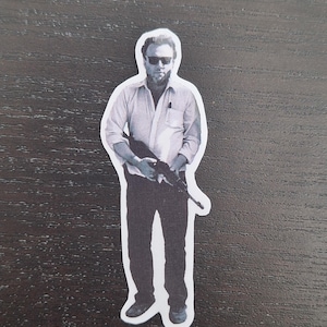 May include: Black and white sticker of a person wearing sunglasses, a button-down shirt, and dark pants, holding a rifle. The sticker has a white border and is placed on a dark wood surface.