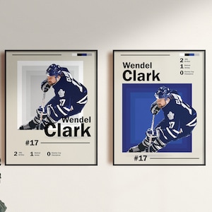 Puede incluir: Dos láminas enmarcadas con un jugador de hockey sobre hielo con uniforme azul y blanco. Las láminas incluyen el nombre "Wendel Clark" y el número "17". Una lámina tiene fondo beige, la otra azul.