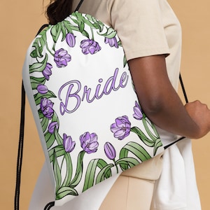 May include: White drawstring bag with a floral design featuring purple tulips and green leaves. The bag has the word "Bride" written in purple script.