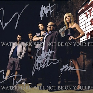 May include: A black and white photo of five people standing in front of a building with a marquee sign that reads "It's Sunny in Philadelphia". The people are all smiling and looking at the camera. The photo is signed by all five people.