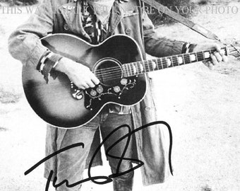 May include: Black and white photo of a man wearing a hat and a long coat, holding a guitar. He is standing in a desert landscape. The photo is signed "Tom Petty".