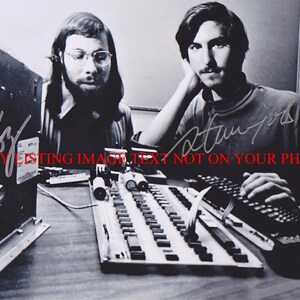 May include: Black and white photo of two men working on a computer. The man on the left is wearing glasses and has a beard. The man on the right is wearing a turtleneck sweater. The photo is signed by both men. The signature on the left says "Woz" and the signature on the right says "Jobs".