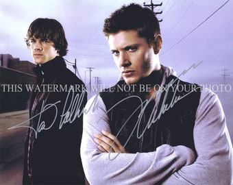 May include: A black and white photo of two men standing side by side. The man on the left is wearing a black jacket and the man on the right is wearing a gray vest over a black shirt. The man on the right has his arms crossed and is looking at the camera. The man on the left is looking off to the side. The photo is signed by both men.
