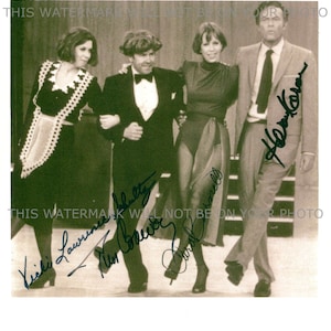 May include: A black and white photo of four people, three women and one man, standing together. The women are wearing dresses and the man is wearing a suit. The photo is signed by all four people. The signatures are "Vicki Lawrence", "Tim Conway", "Carol Burnett", and "Harvey Korman".