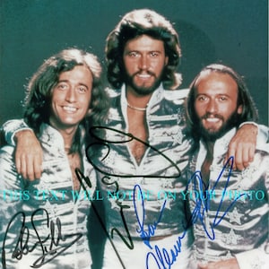 May include: A black and white photo of three men in matching white and silver outfits with a sparkly design. The men are smiling and looking at the camera. The photo is signed by each of the men. The signatures are in black ink.