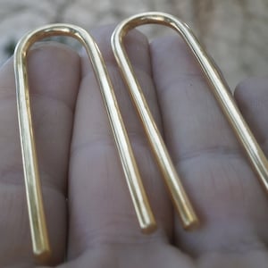 Peut inclure: Une paire de boucles d'oreilles minimalistes de couleur or avec un design simple et incurvé. Les boucles d'oreilles sont faites d'un métal fin et lisse et ont une forme légèrement arrondie.