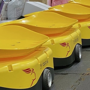 Puede incluir: Una fila de cuatro vehículos de guiado automático (AGV) amarillos con un logotipo de alas rojas y el texto "100%16" en el lateral. Los AGV se utilizan para transportar mercancías en un almacén o fábrica.