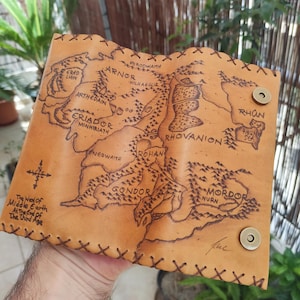May include: A tan leather wallet featuring a detailed map of Middle Earth, with regions like Mordor and Gondor. The map is etched into the leather, and the wallet has a stitched border and magnetic snap closures. Text on the wallet reads "The West of Middle Earth At The End of The Third Age."